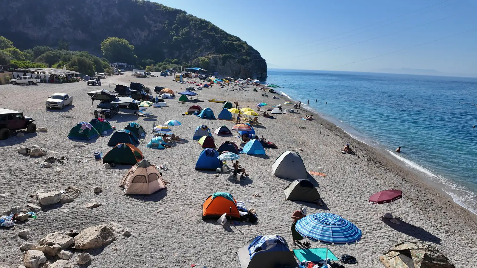 Tents and sun umbrellas across Gjipe beach under a tall cliff, with the sea on the right