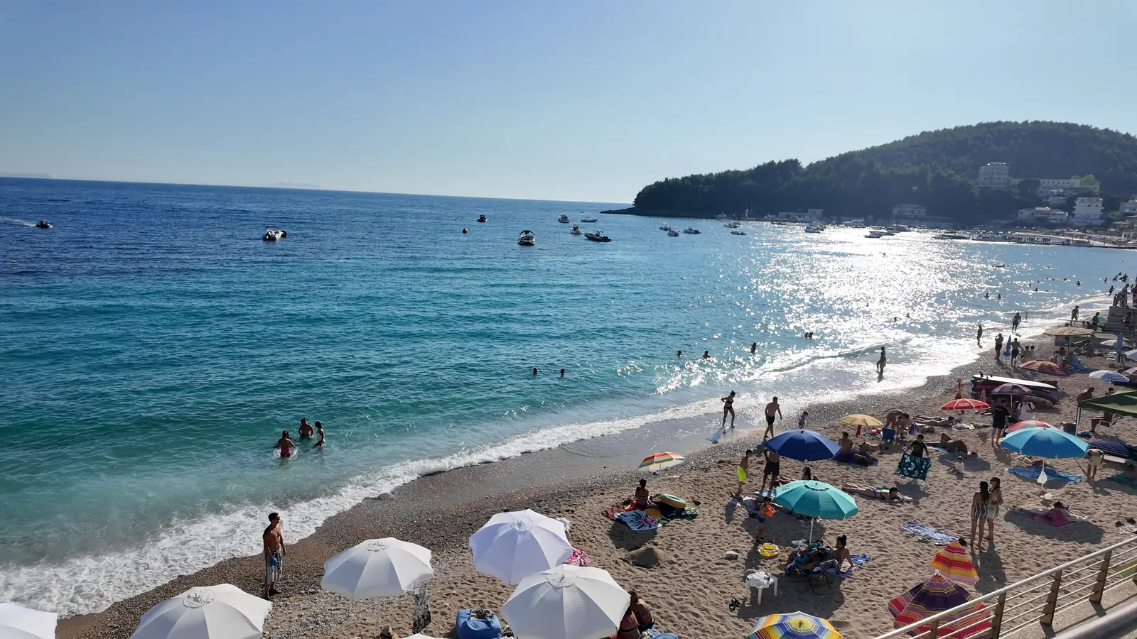 The long pebble beach in Himarë with white umbrellas, clear water and the green headland behind