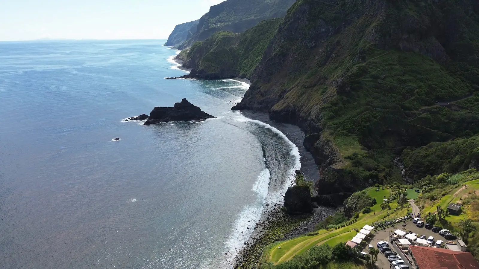 Green sea cliffs dropping to a thin black beach on the coast of Madeira, seen from the air
