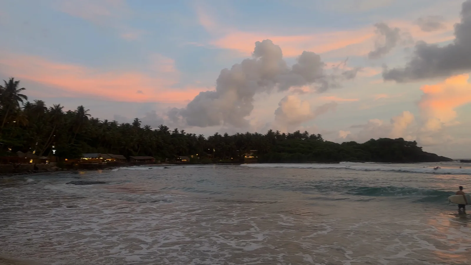 Hiriketiya bay at dusk under pink clouds, with a surfer waiting in the water