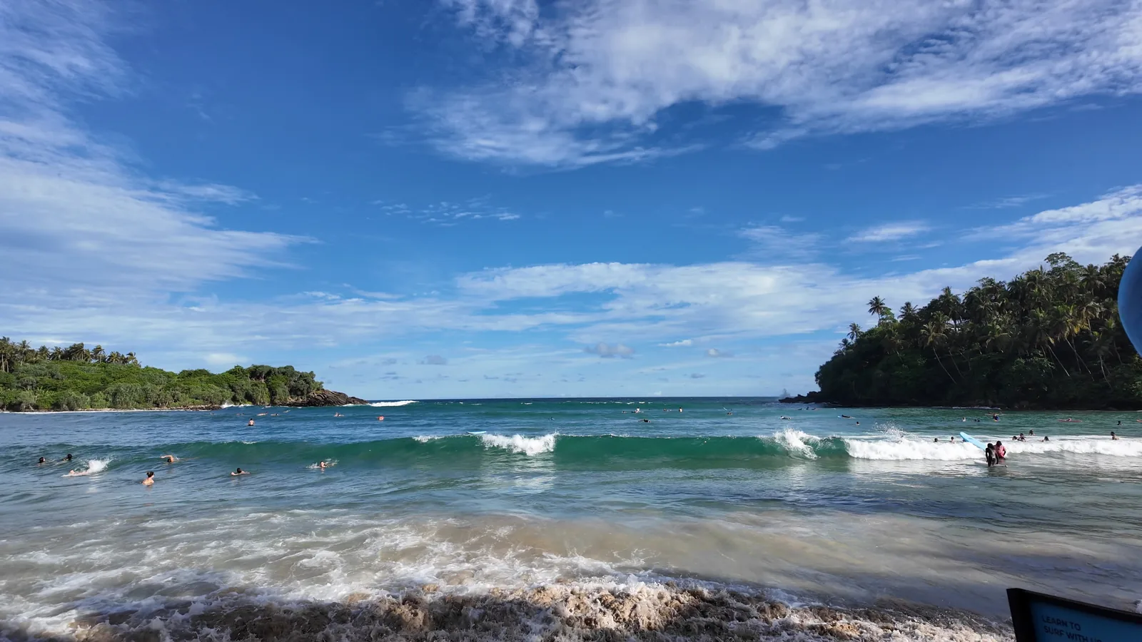 Waves rolling into Hiriketiya beach under a big blue sky, with the green headland on the left